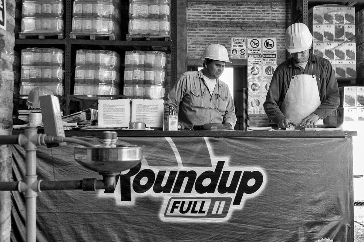 Two workers in hard hats at a counter draped with a Roundup banner, with shelves of product containers behind them.