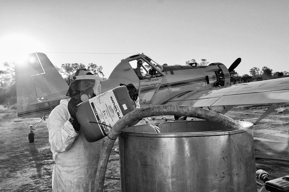 A technician in protective clothing and a respirator pours a chemical container into a tank beside a crop-dusting aircraft.