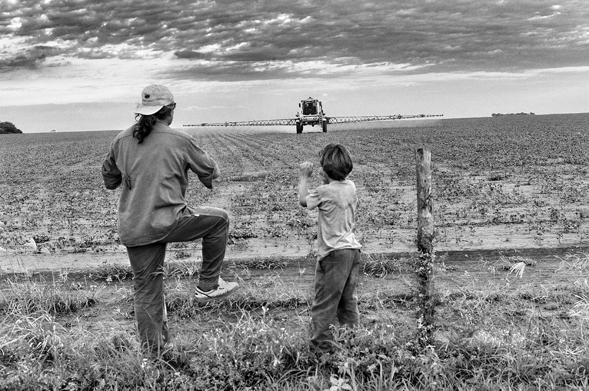A man and a boy stand at a wire fence watching a sprayer cross a large field.