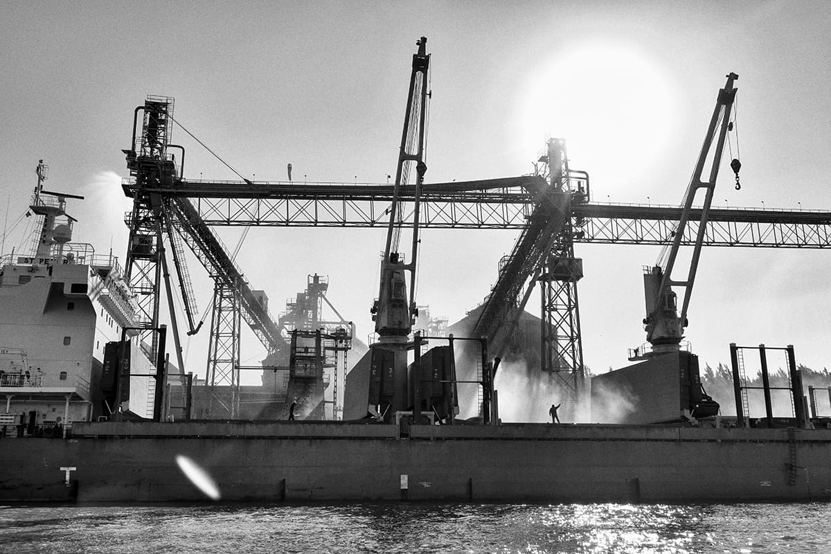 Loading cranes above a cargo ship at a river port, backlit by the sun.