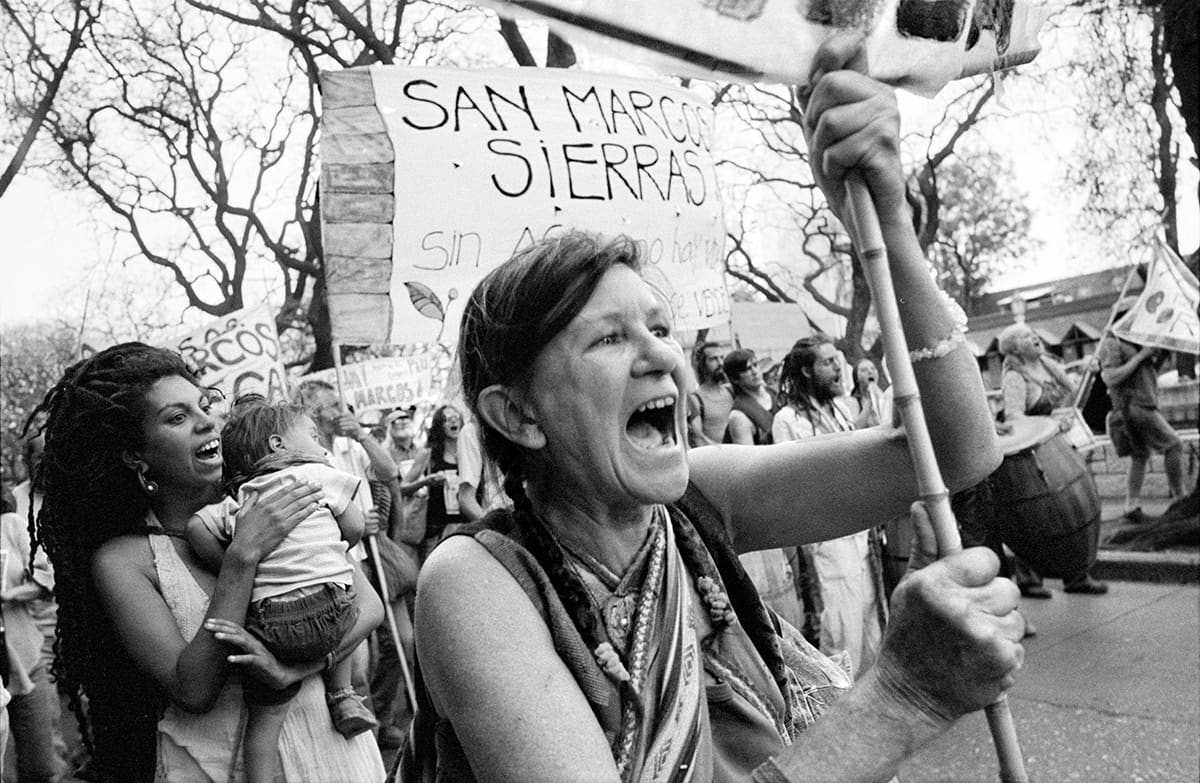 Women shout during a street march, one holding a child, with protest signs behind them.