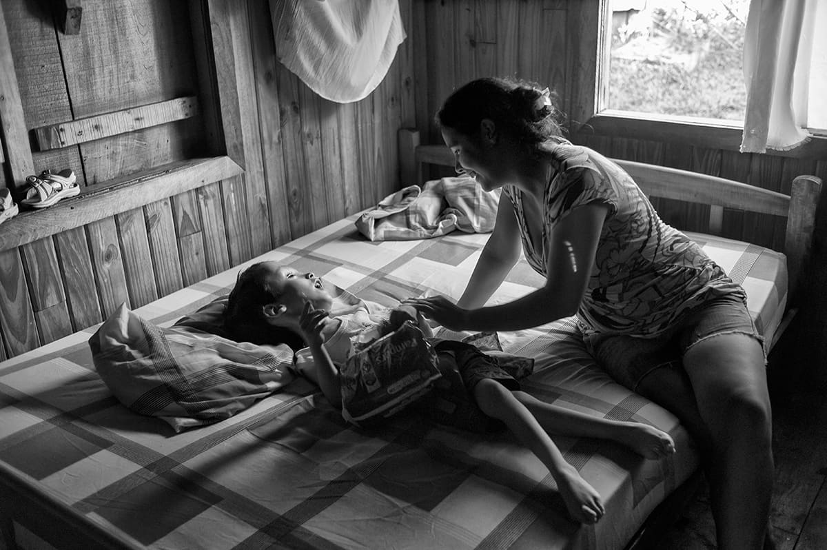 A mother leans over her son lying on a bed in a wooden room.