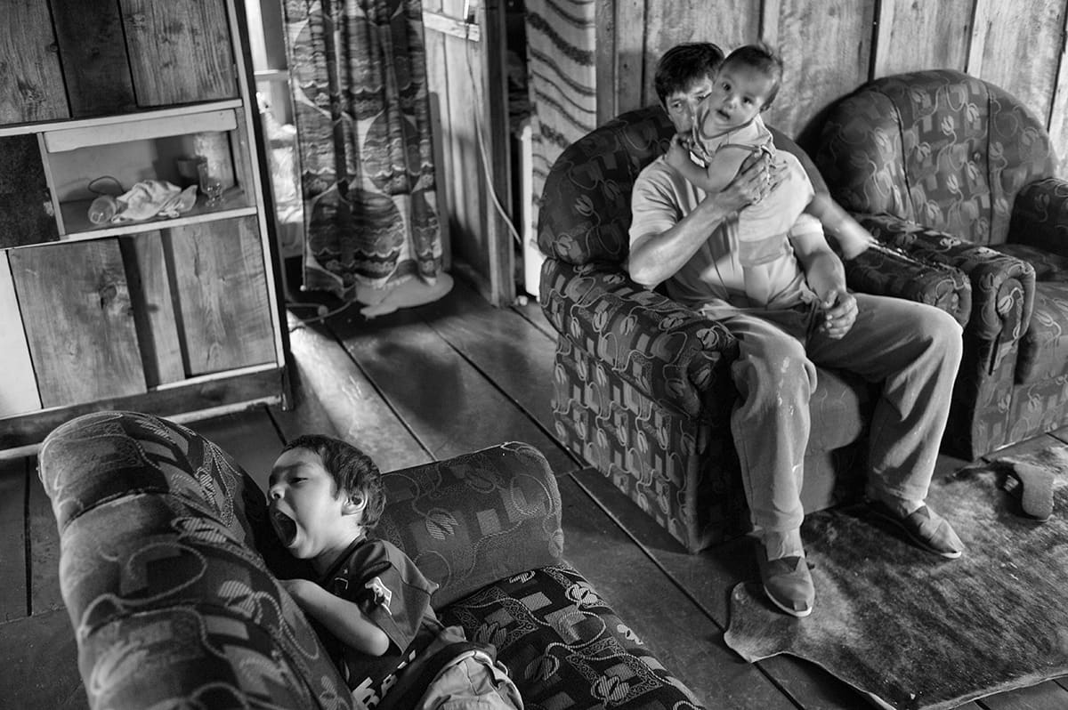 A man holds a young child on an armchair in a living room while another child yawns on a sofa.