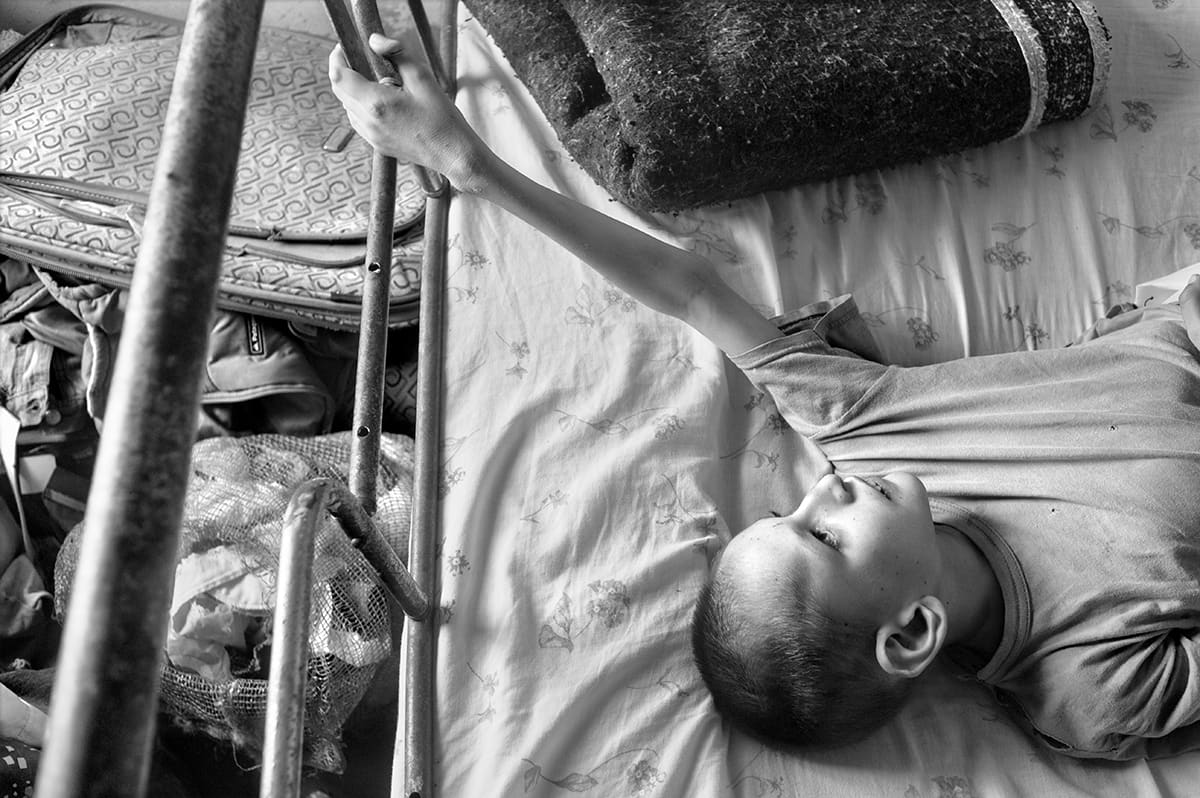 A boy lies on his back on a bed, one arm raised to grip the metal bed frame.