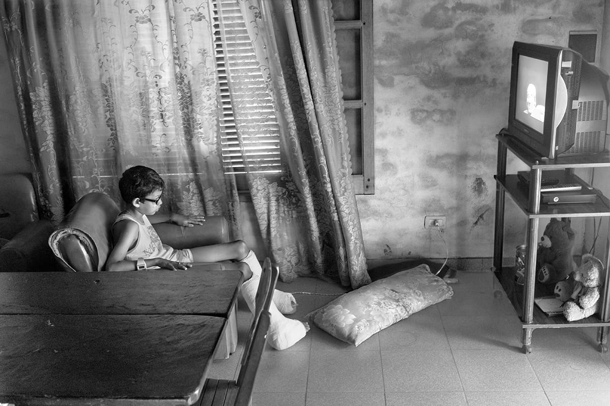 A boy in glasses sits in an armchair in a living room beside a curtained window and a television.
