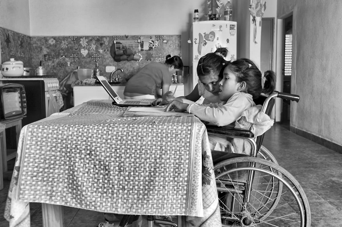 A girl in a wheelchair sits at a kitchen table, embraced by her sister, while a woman works at the stove.