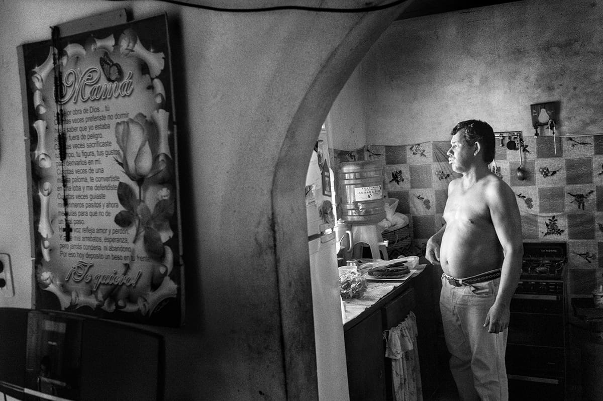 A shirtless man stands in a kitchen doorway beside a framed prayer on the wall.