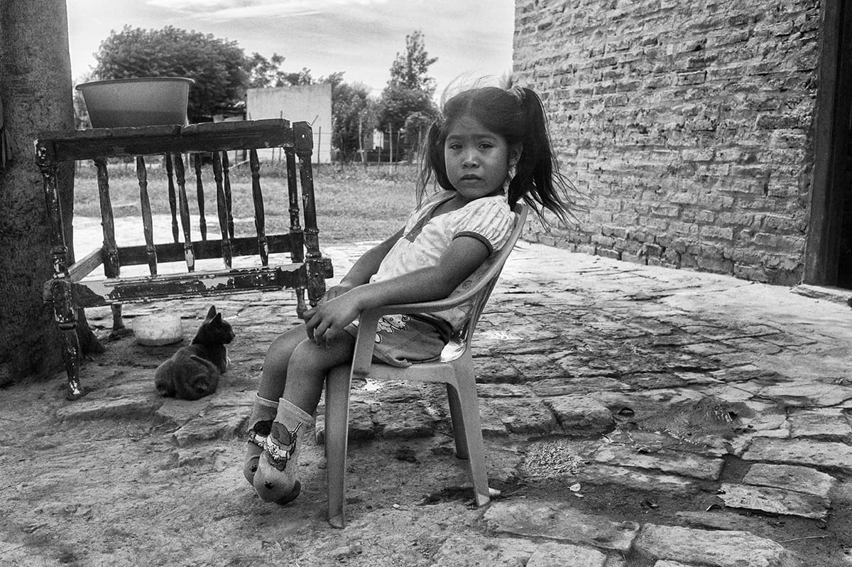 A young girl sits on a plastic chair in a paved courtyard, a cat resting nearby.