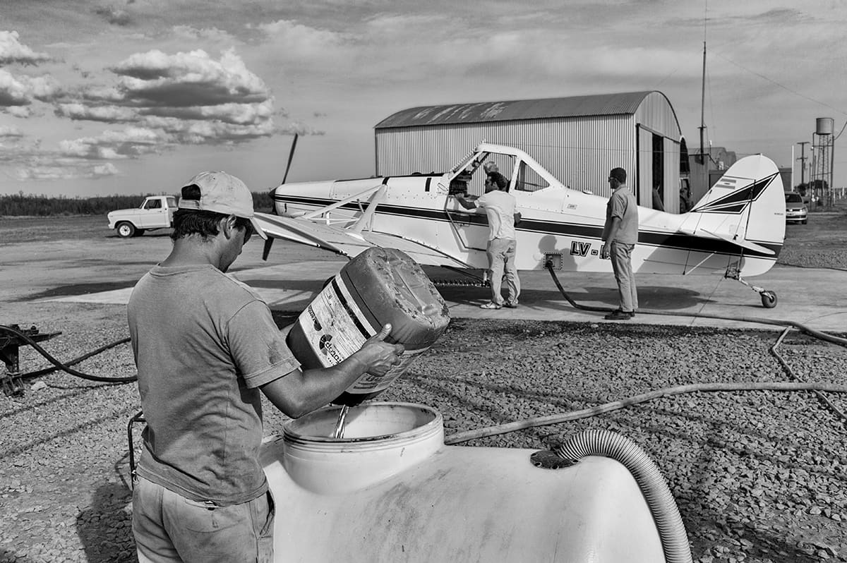 A man pours a chemical container into a tank beside a small crop-dusting aircraft on an airstrip.