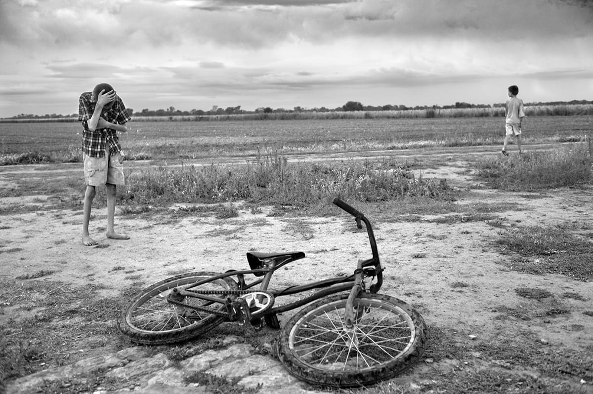 A boy covers his face with his hand beside a bicycle lying on the ground at the edge of a field.