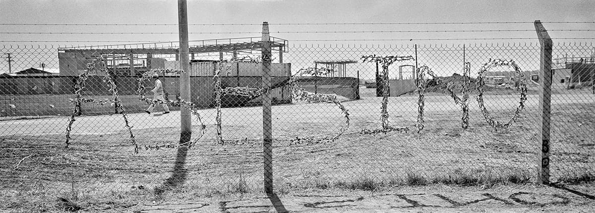 A word shaped from wire hangs on a chain-link fence in front of an industrial site.
