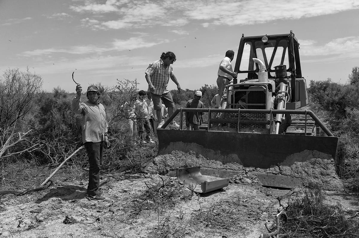 A group of men stand on and around a bulldozer in dry scrubland, one raising a machete.