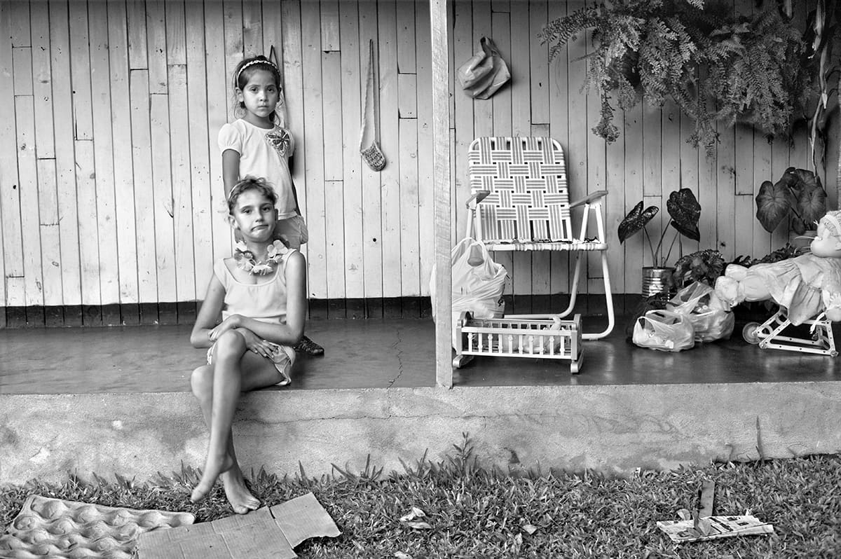 Two girls on the porch of a wooden house, one sitting on the step, the other standing behind her.