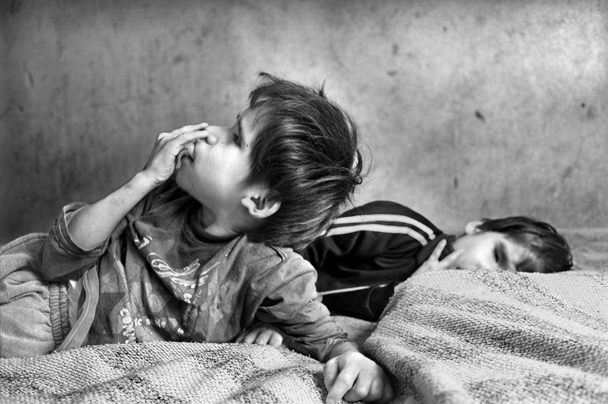 Two boys lie on a bed under a blanket, one propped on his elbow with a hand over his face.