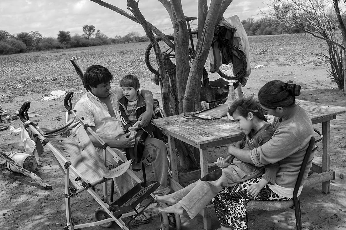 A family sits at a wooden table under a tree outside their home, holding two young boys.