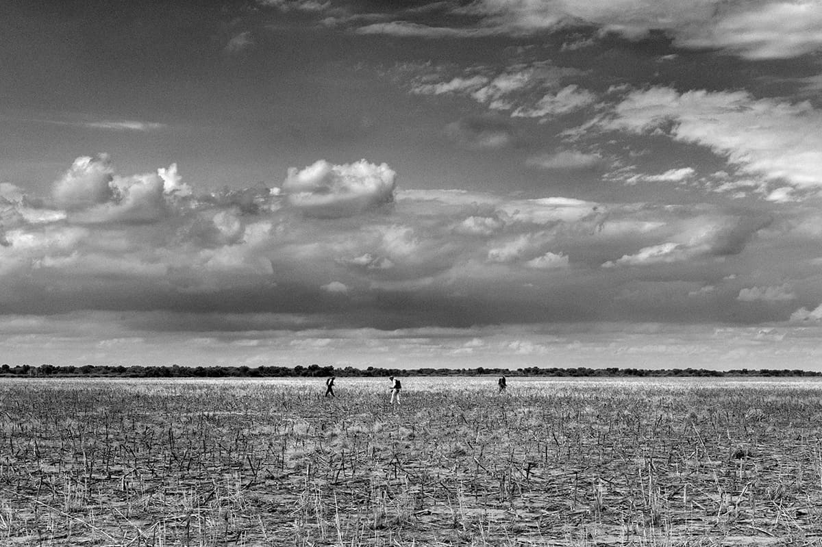 Three small figures cross a vast flat field under a wide cloudy sky.