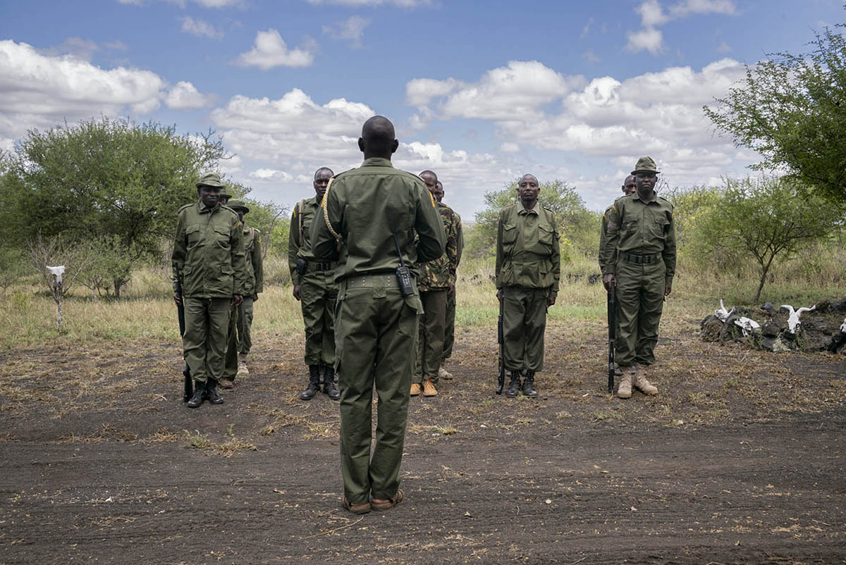 Uniformed rangers stand in a line on bare ground facing their captain, seen from behind.