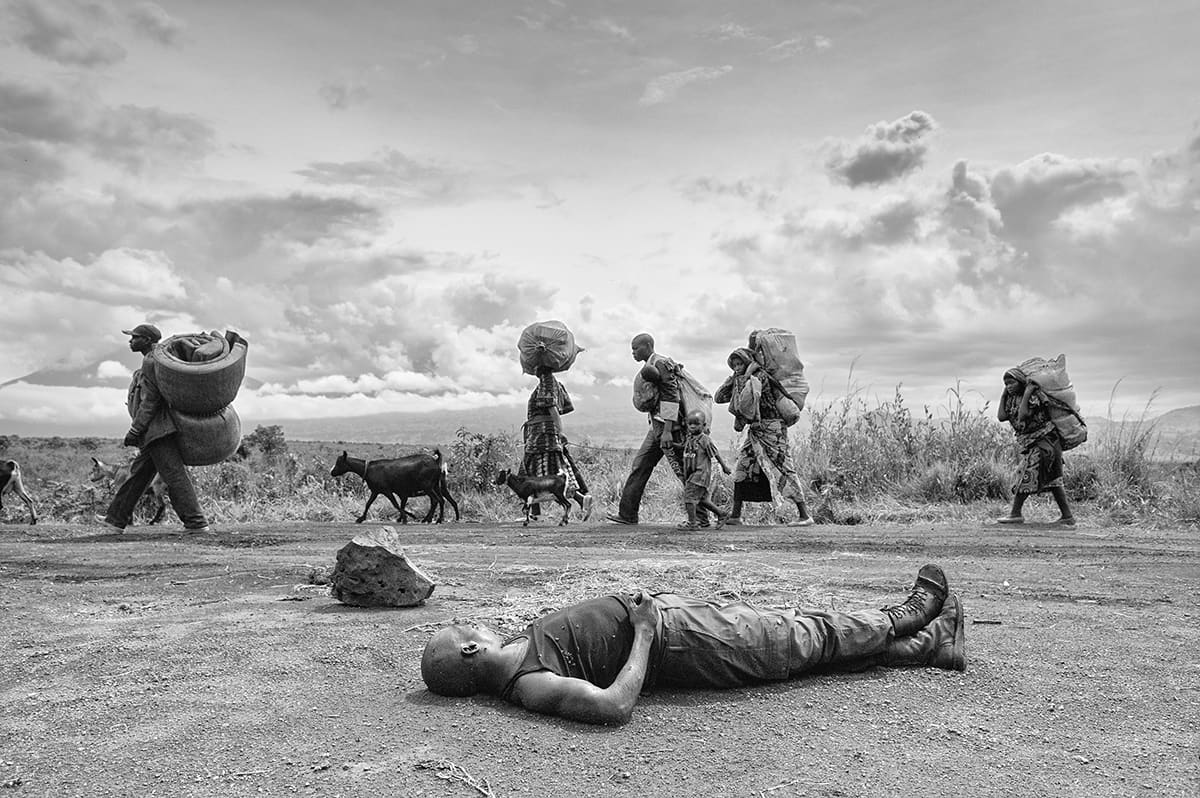 The body of a dead Congolese army soldier on a road as civilians flee past with their belongings.