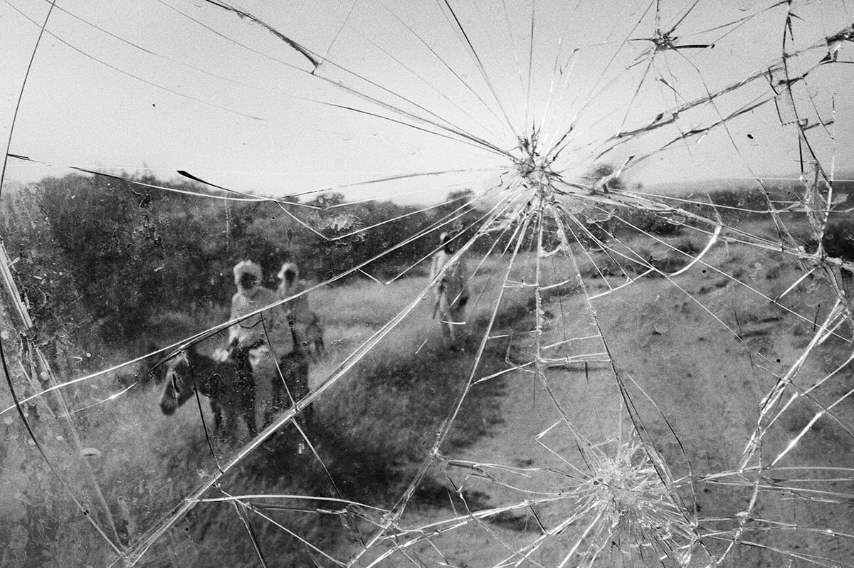 Displaced family walking with a donkey seen through the shattered windscreen of a vehicle in Darfur.