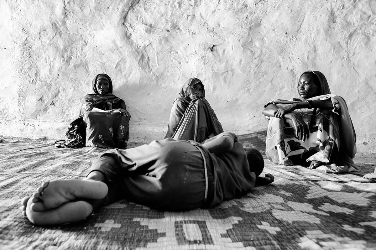 Displaced women and a sleeping child resting on a mat inside a shelter in Darfur, Sudan.