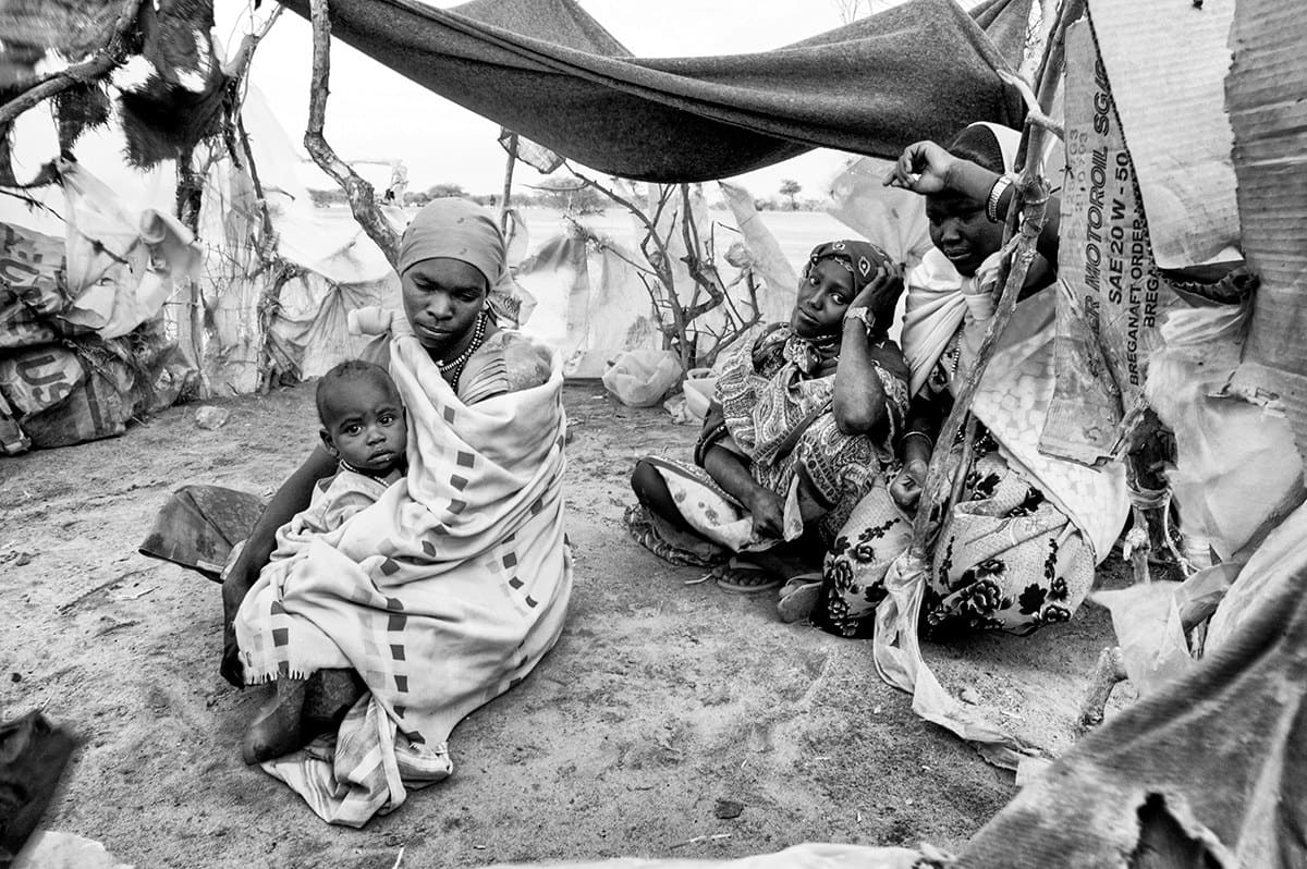 Displaced women and a young child sheltering under blankets and plastic sheeting at a camp in Darfur, Sudan.