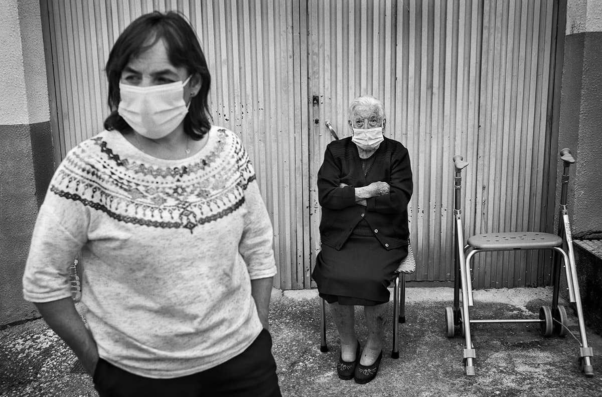 Elderly woman wearing a face mask sitting beside her walking frame while a masked woman stands in the foreground in a Spanish village.