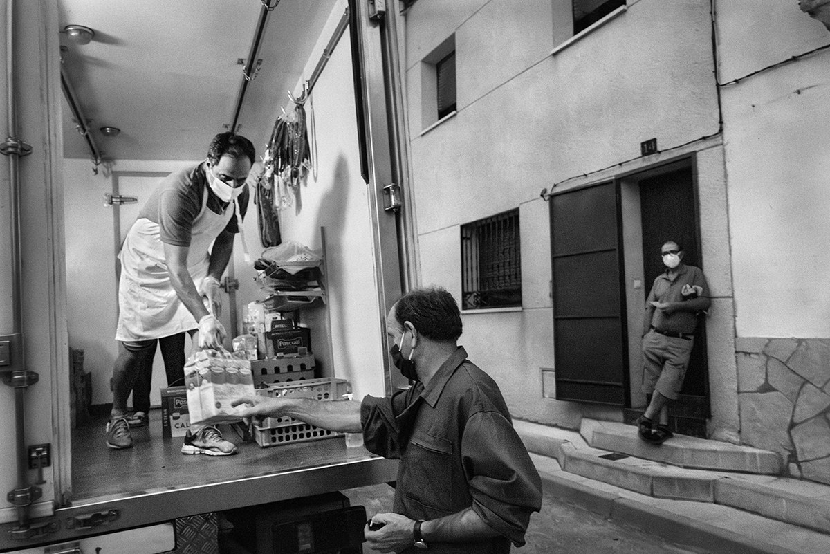Volunteer handing out food supplies from a truck to a masked resident in a narrow street in Spain during the pandemic.