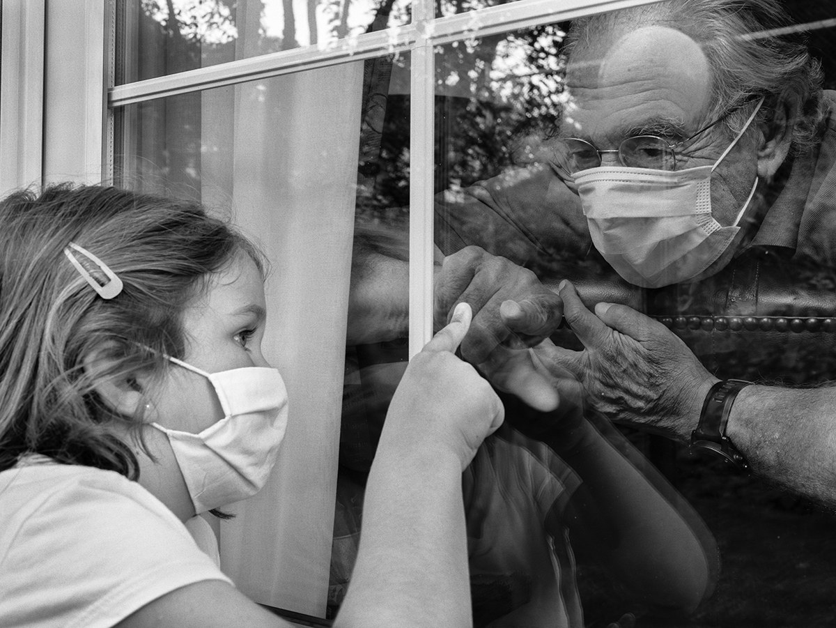 Girl wearing a face mask touching a window while her grandfather looks at her from the other side of the glass during the COVID-19 pandemic in Spain.