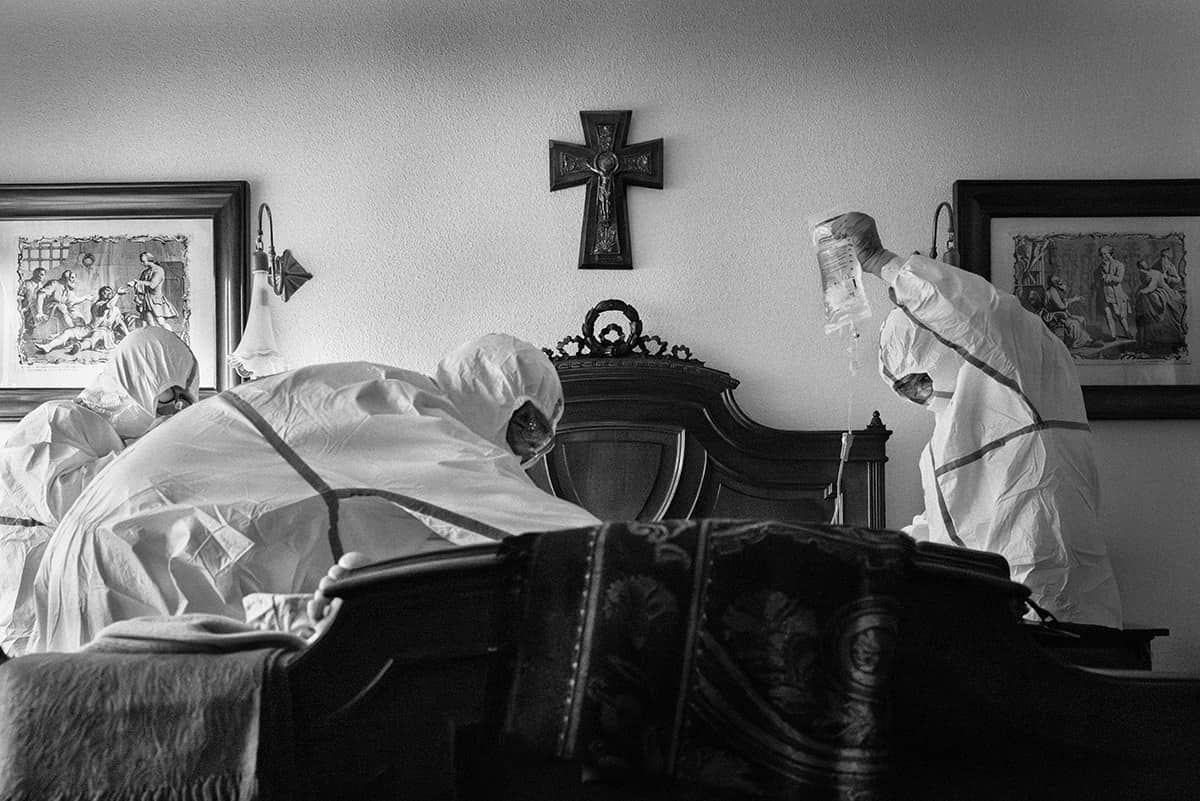 Medical team attending a COVID-19 patient at home in a bedroom with a crucifix on the wall in Spain.