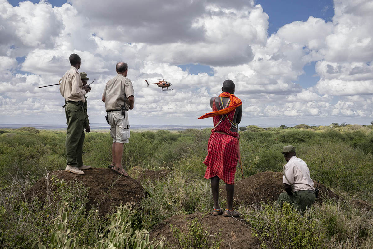 Men standing on mounds in the bush watch a small helicopter in the sky; one wears a red checked shuka.