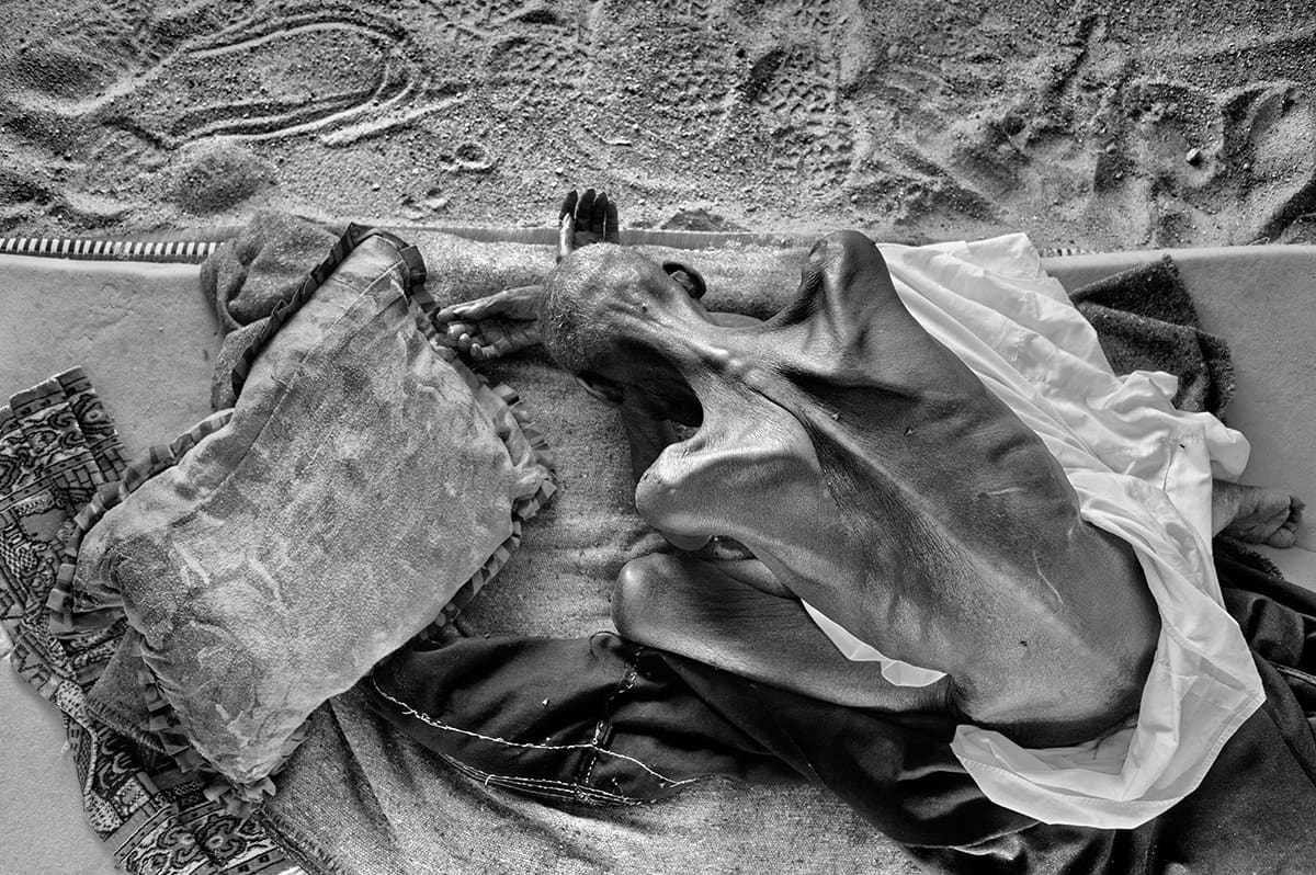 Severely malnourished man lying on a mat on the sand at a camp in Darfur, Sudan.