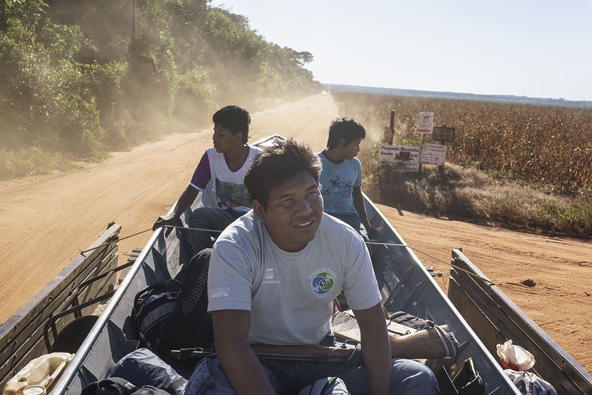 Manoki men traveling on a boat-shaped vehicle along a dirt road bordering plantations