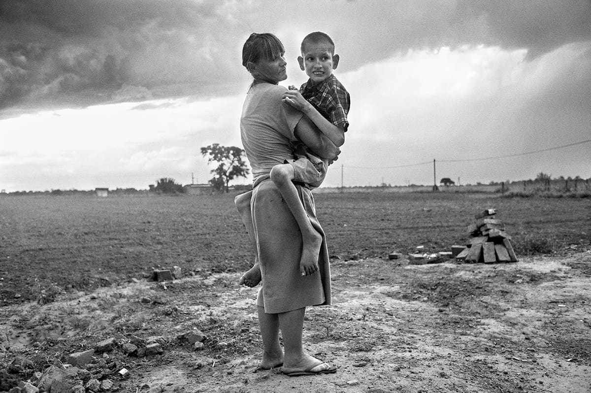 A woman holds a boy in her arms on bare ground beside a flat cultivated field under a stormy sky.