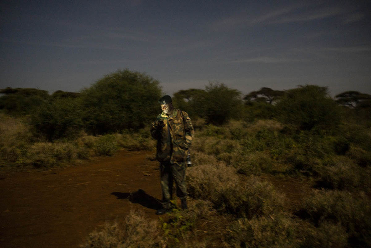 A ranger stands alone on a dirt track in the dark, his face lit by the glow of a mobile phone.