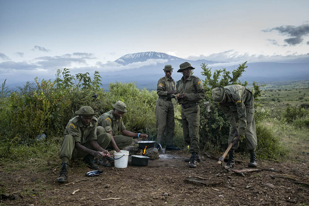 Rangers prepare and eat a meal at a hillside camp, with Kilimanjaro rising above the clouds behind them.