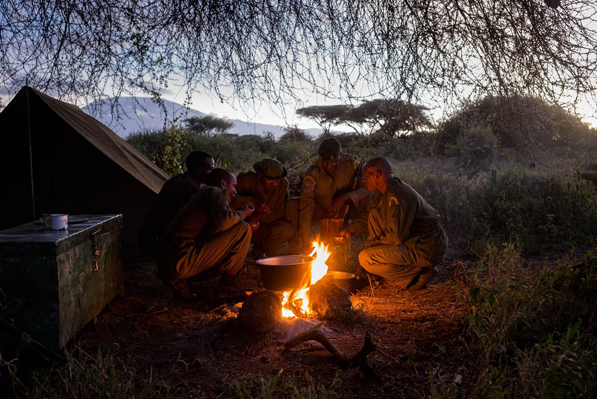 Rangers crouch around a cooking fire beside a tent at dawn, with Kilimanjaro faint in the distance.