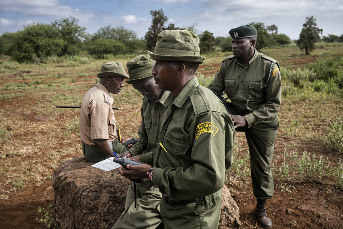 Four uniformed men gather around a rock, one writing in a notebook while the others look on.
