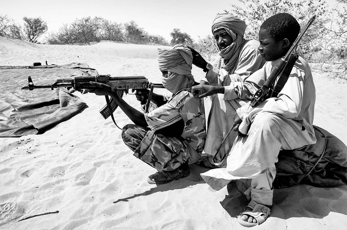 Darfur rebel fighters aiming an AK-47 rifle from the sand during a firefight in Sudan.