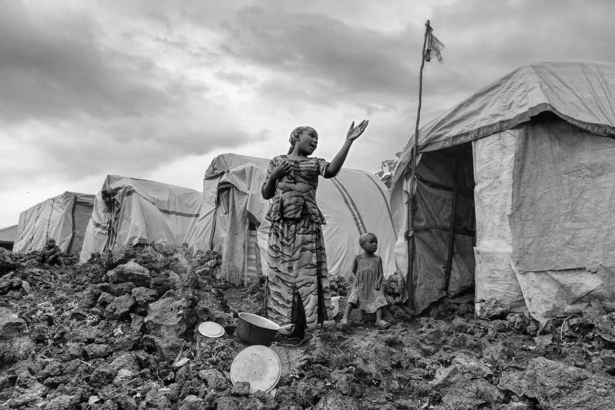 A woman standing among the tents of the Kibati refugee camp with a child sitting on a rock.