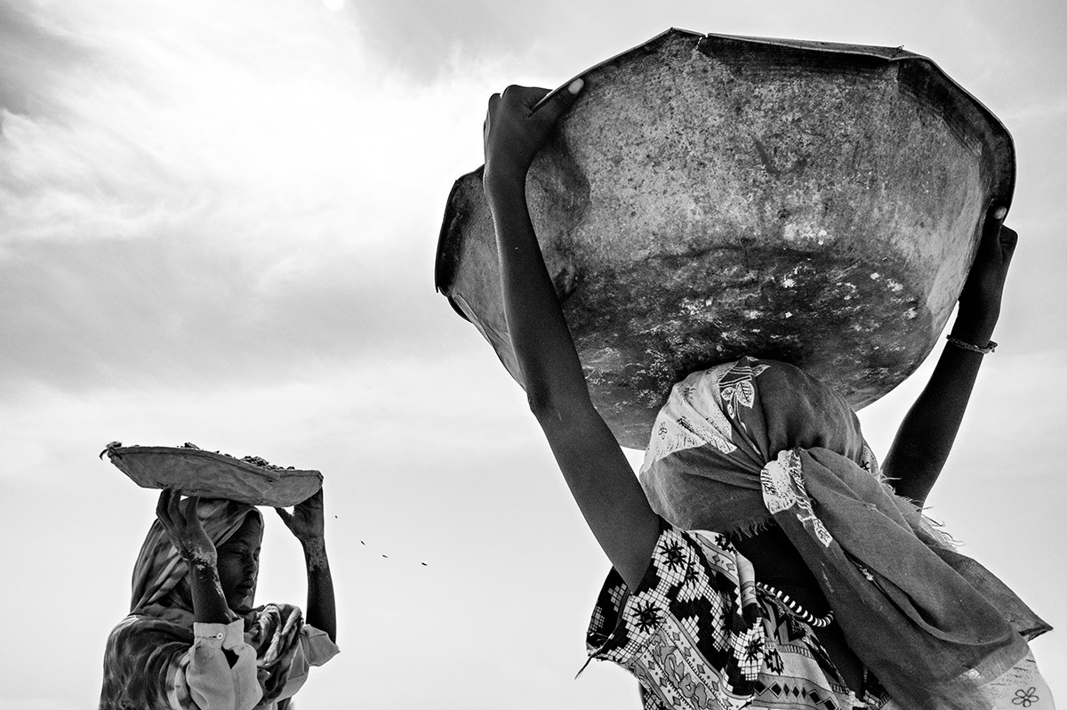 Two Darfuri women carrying large metal bowls on their heads against the sky in Sudan.